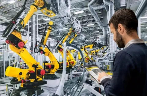 Ein Ingenieur verwaltet mit einem Tablet eine Fabrikhalle voller gelber FANUC-Roboterarme.